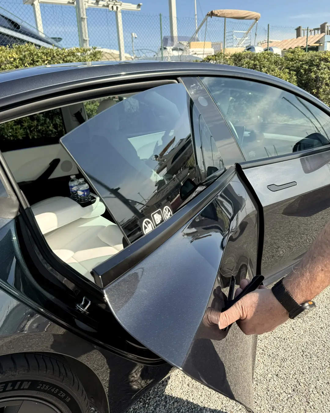 Une main ouvre la portière arrière de la Tesla Model 3 ELYSIA sur la banquette blanche et deux bouteilles d’eau, au port de Saint-Laurent-du-Var